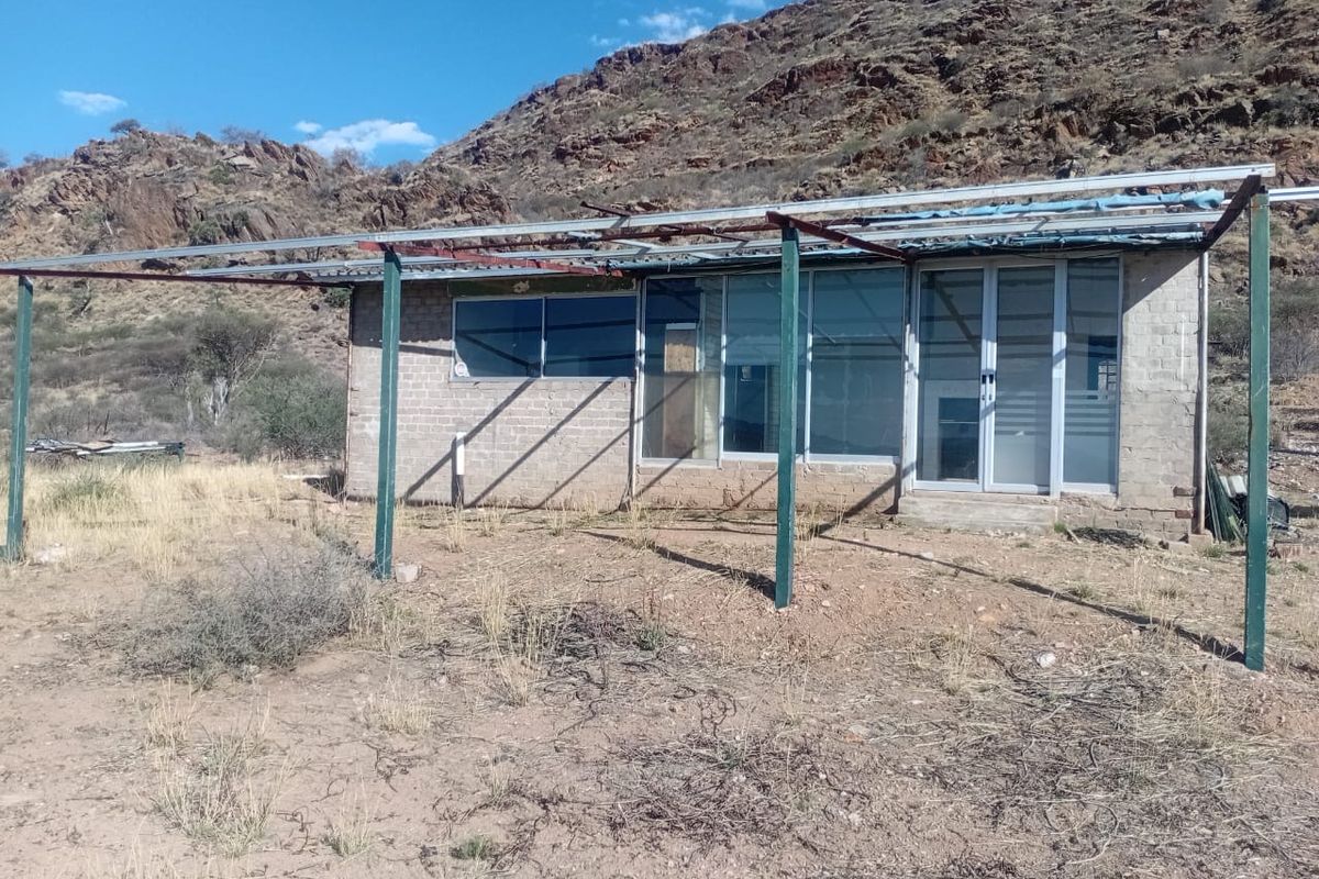 Rear view of a small structure featuring a metal pergola frame, sliding glass doors, and aluminum window frames set against a rocky hillside.
