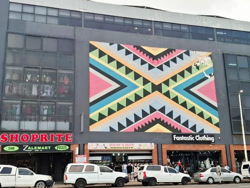 Front elevation of a commercial retail center featuring multiple storefronts, large glass windows, and a prominent decorative mural on the facade.