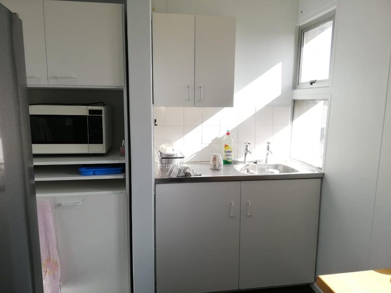 A compact office kitchenette featuring white built-in cabinetry, a stainless steel sink, tiled splashback, and natural light from adjacent windows.