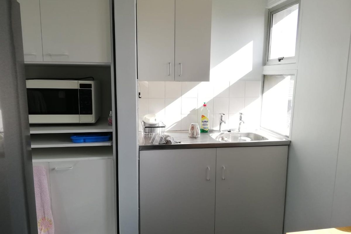 A compact office kitchenette featuring white built-in cabinetry, a stainless steel sink, tiled splashback, and natural light from adjacent windows.