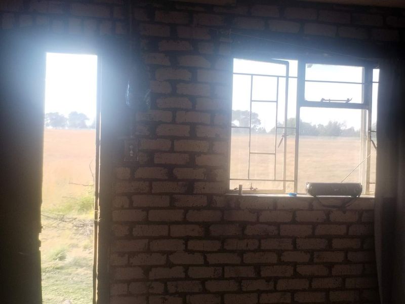 A rustic, unfinished room featuring exposed brick walls and steel-framed windows with security bars, offering a view of the surrounding farm landscape.