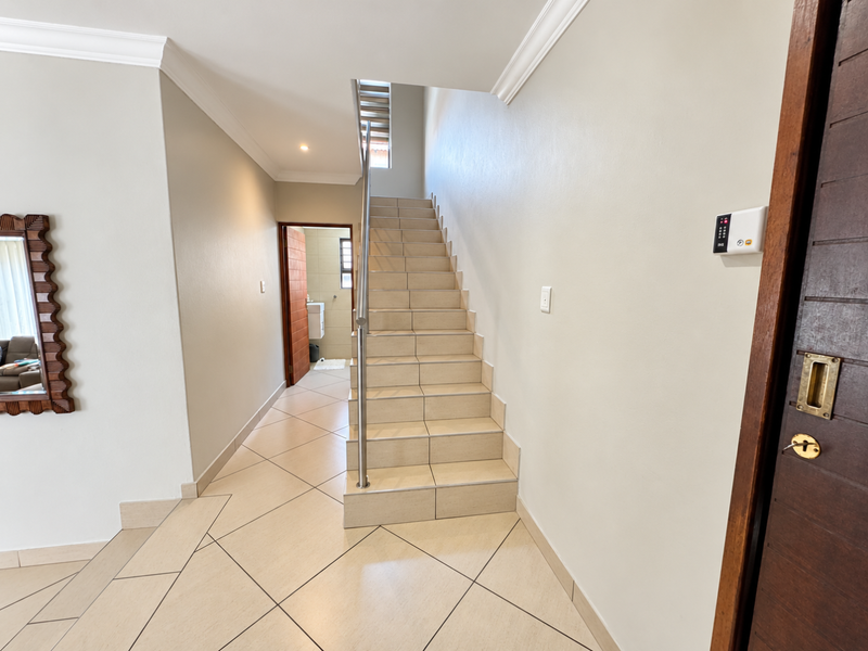 A bright, tiled hallway featuring a staircase with a stainless steel handrail, crown molding, and a secure entrance door.