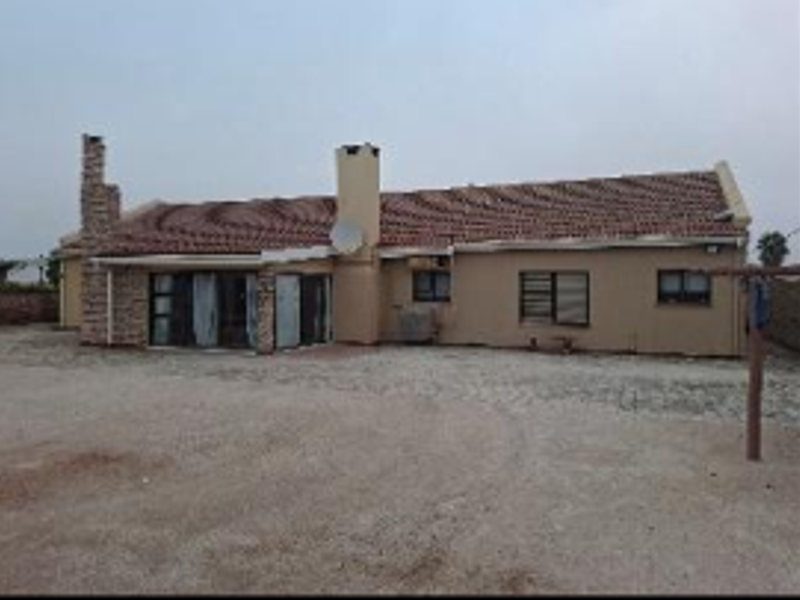 Rear view of a single-story house featuring a tiled roof, stone chimney, and sliding glass doors opening to an unpaved yard area.