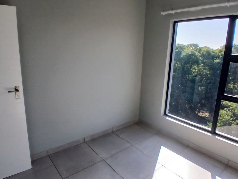 A bright, empty bedroom featuring neutral tiled flooring, a large window with natural light, and a clean, modern interior finish.