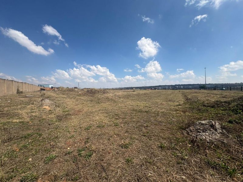 A wide-angle view of a vacant commercial land plot featuring dry grass, uneven terrain, and a perimeter boundary wall.