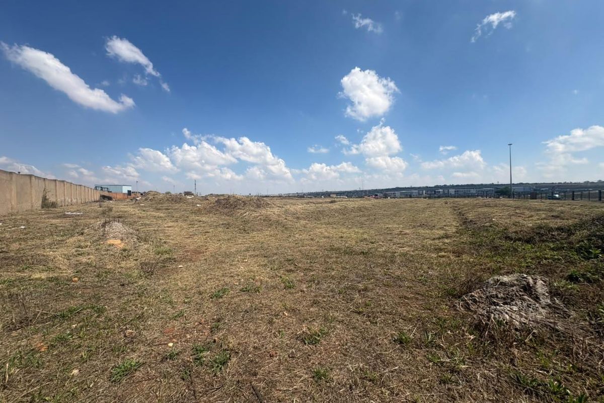 A wide-angle view of a vacant commercial land plot featuring dry grass, uneven terrain, and a perimeter boundary wall.