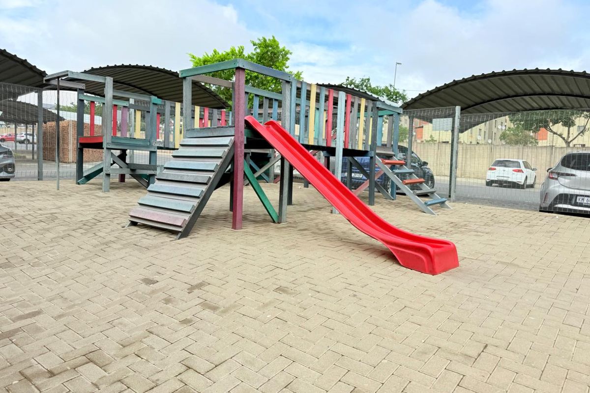 Outdoor communal playground area featuring a multi-level wooden climbing structure with a slide, situated on paved ground within a residential complex.