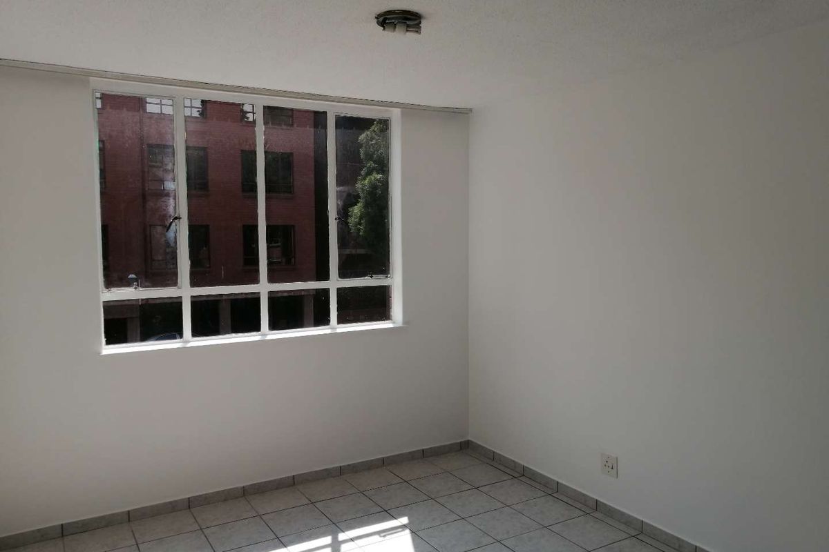 A bright, empty bedroom featuring tiled flooring, a large window with an urban view, and a simple ceiling light fixture.