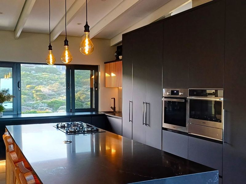 Modern kitchen featuring dark cabinetry, a large central island with a gas hob, built-in ovens, and expansive windows with scenic views.