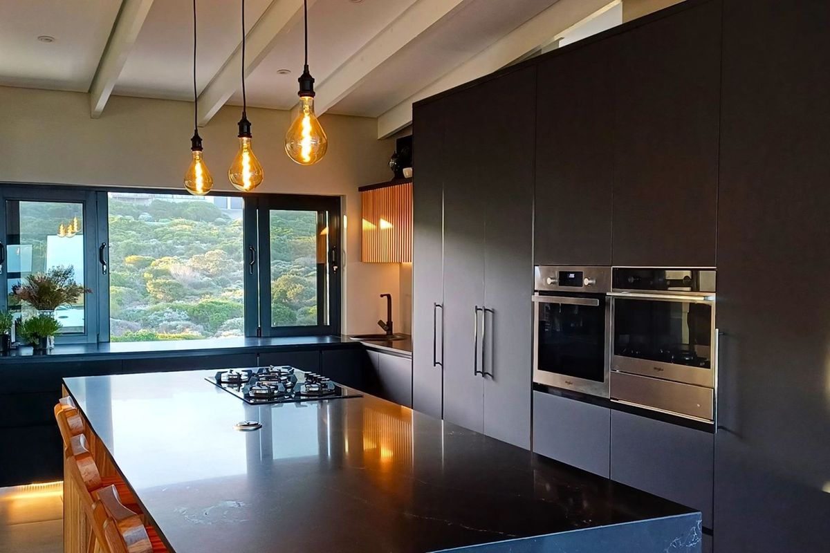 Modern kitchen featuring dark cabinetry, a large central island with a gas hob, built-in ovens, and expansive windows with scenic views.