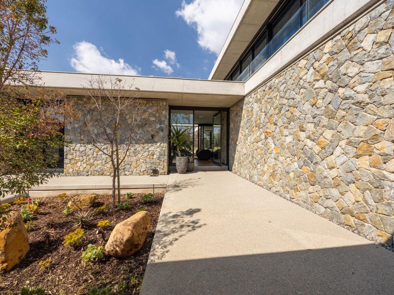 Modern architectural entrance featuring natural stone cladding, a clean concrete walkway, and expansive glass doors leading into the home.
