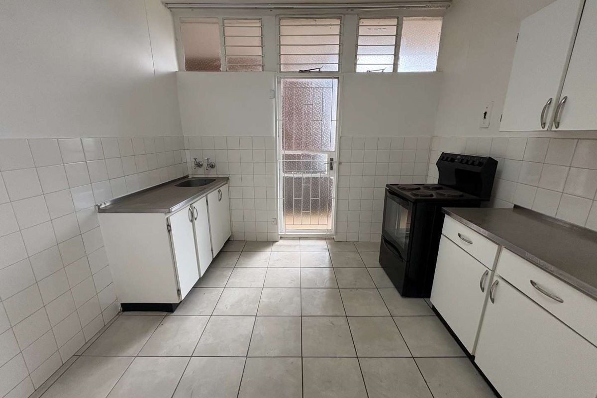 A functional, empty kitchen featuring tiled flooring, white cabinetry, a stainless steel sink, and a freestanding electric stove with access to an exterior door.