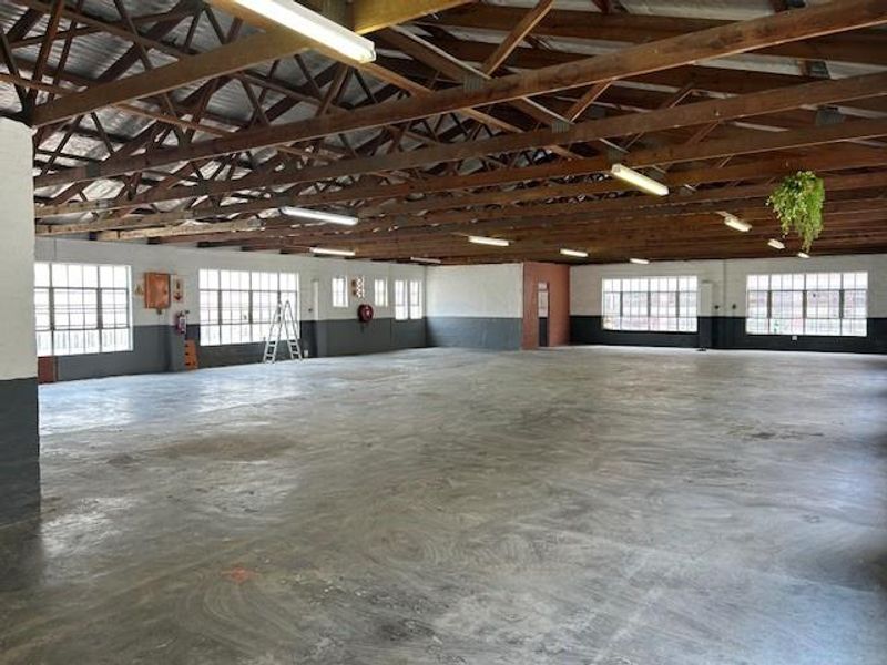 Spacious industrial warehouse or utility area featuring exposed timber roof trusses, concrete flooring, and large steel-framed windows providing ample natural light.