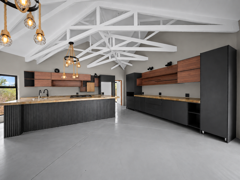 Modern kitchen featuring exposed white roof trusses, sleek dark cabinetry with wood accents, a large island, and polished concrete floors in an open-plan layout.