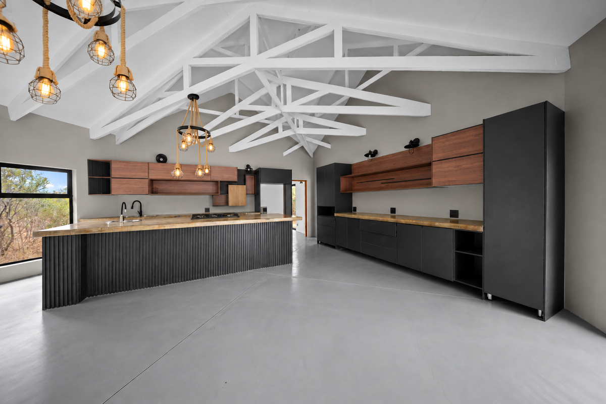 Modern kitchen featuring exposed white roof trusses, sleek dark cabinetry with wood accents, a large island, and polished concrete floors in an open-plan layout.