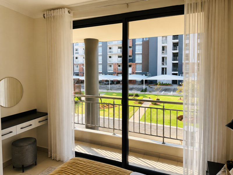 A bright bedroom featuring a built-in vanity and a sliding glass door that opens onto a private balcony with views of the complex grounds.