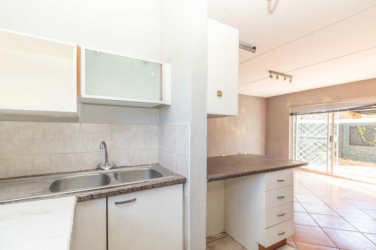 A dated kitchen area featuring a double sink, laminate countertops, and built-in cabinetry, opening into a living space with tiled flooring.