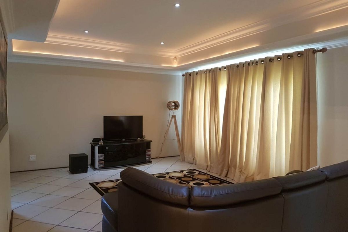 A comfortable living room featuring tiled flooring, a recessed ceiling with integrated cove lighting, and large window treatments.