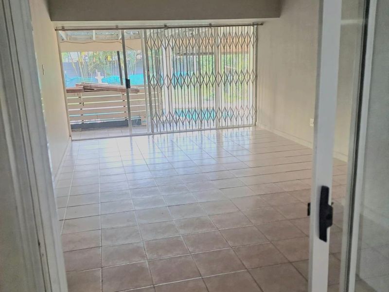 An empty, tiled living room featuring large sliding glass doors with a security gate, providing direct access to an outdoor patio area.