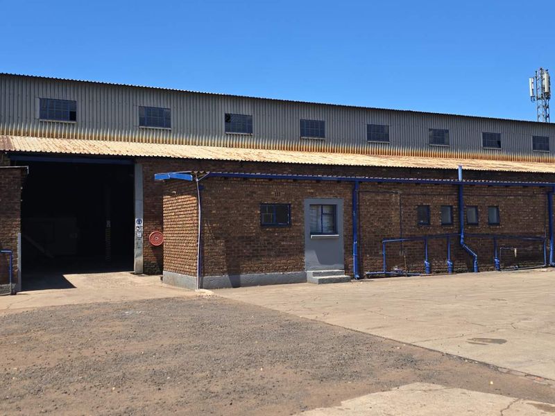 Industrial warehouse exterior featuring exposed brickwork, corrugated metal roofing, and a large roller shutter door access point with an expansive concrete yard.