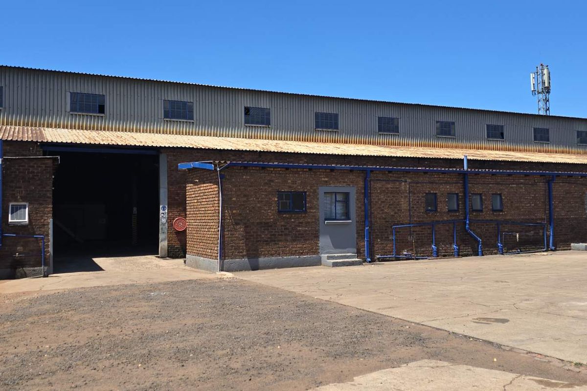 Industrial warehouse exterior featuring exposed brickwork, corrugated metal roofing, and a large roller shutter door access point with an expansive concrete yard.