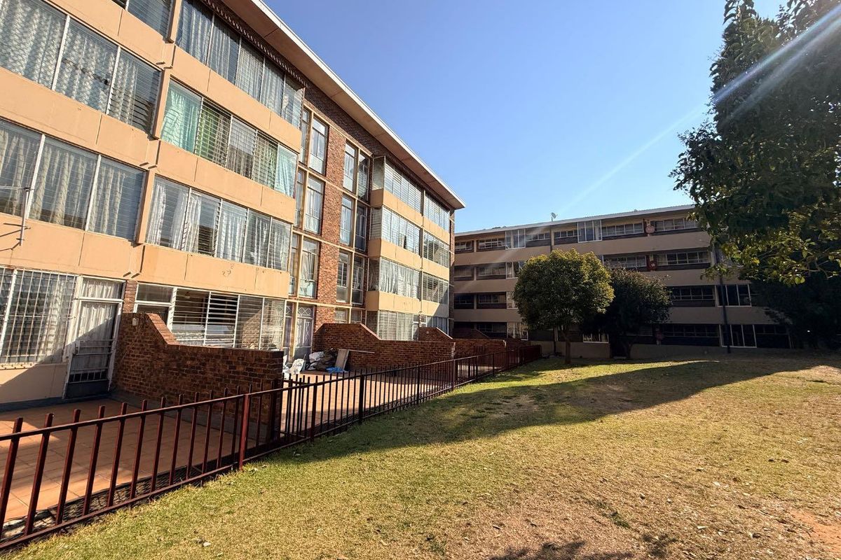 Exterior view of an apartment complex featuring brick facades, ground floor patios with security fencing, and a shared communal garden area.