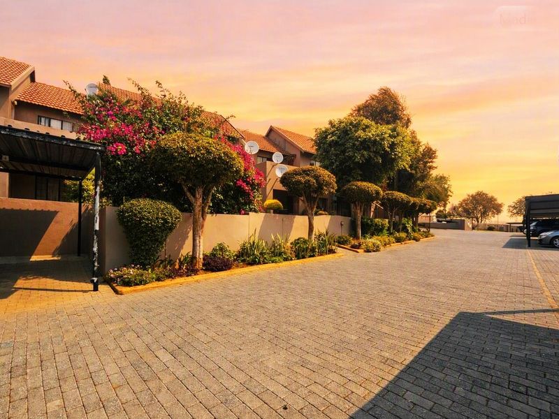 A well-maintained apartment complex exterior featuring a paved driveway, covered carport parking, and established landscaped garden beds along the boundary wall.