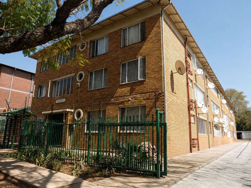Exterior view of a multi-story face brick apartment building featuring secure perimeter fencing, paved access, and multiple satellite dish installations.
