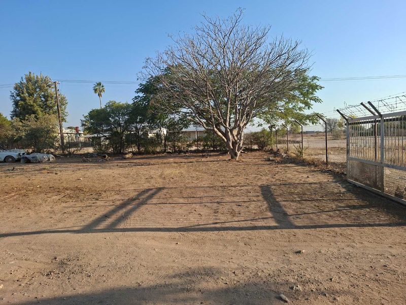An expansive, undeveloped rear outdoor area featuring security fencing, a metal gate, and mature trees on a dirt surface.