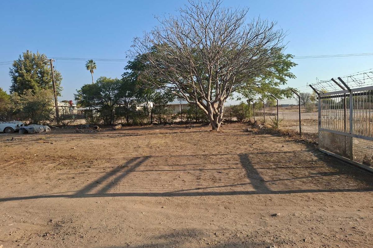 An expansive, undeveloped rear outdoor area featuring security fencing, a metal gate, and mature trees on a dirt surface.