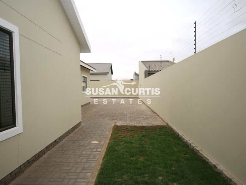 A side view of the property featuring a paved walkway, a small lawn area, and secure boundary walls with electric fencing.
