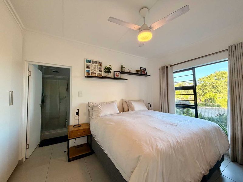 A well-lit bedroom featuring tiled flooring, a ceiling fan, and an en-suite shower room with large windows providing natural light.