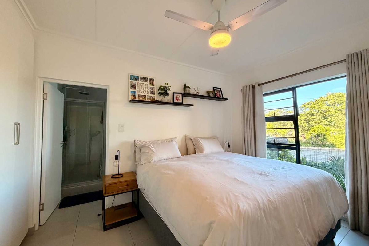 A well-lit bedroom featuring tiled flooring, a ceiling fan, and an en-suite shower room with large windows providing natural light.