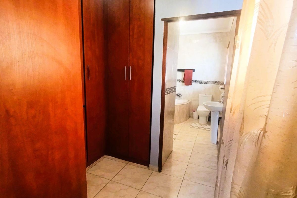 A view from a dressing area with built-in wooden cupboards leading into a functional bathroom featuring a pedestal basin and corner bathtub.