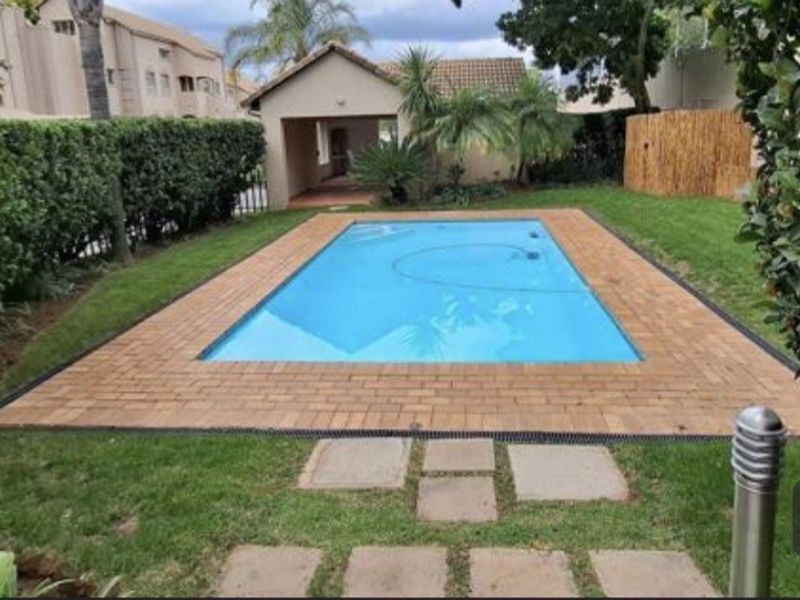A well-maintained swimming pool area featuring brick paving, surrounding lawn, and established garden hedging.
