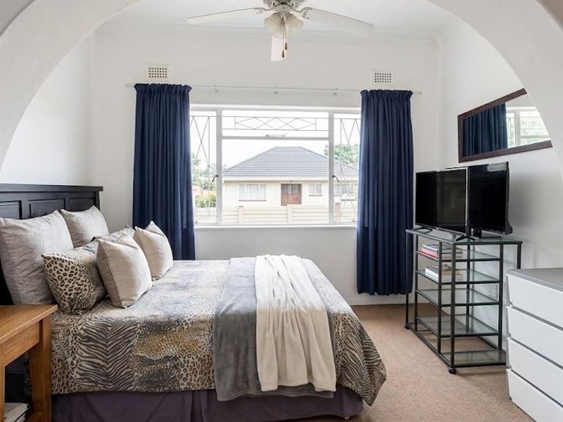 A well-lit bedroom featuring a classic arched entryway, ceiling fan, and large window with security bars, offering a comfortable and functional living space.