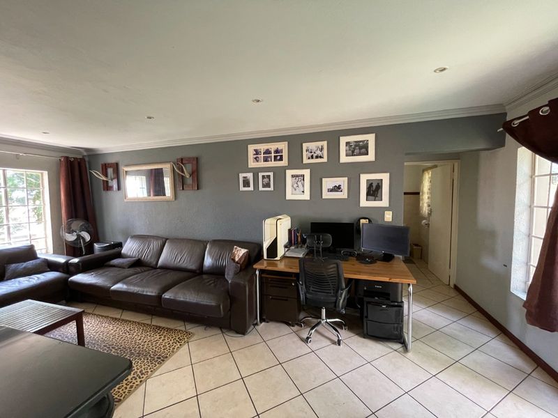 A spacious living room featuring neutral ceramic floor tiles, recessed downlighting, and a neutral grey feature wall, offering a versatile layout for relaxation and work.