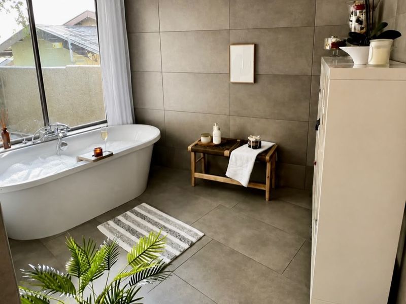 A modern, well-appointed bathroom featuring a sleek freestanding bathtub, large-format neutral tiling, and a large window providing natural light.