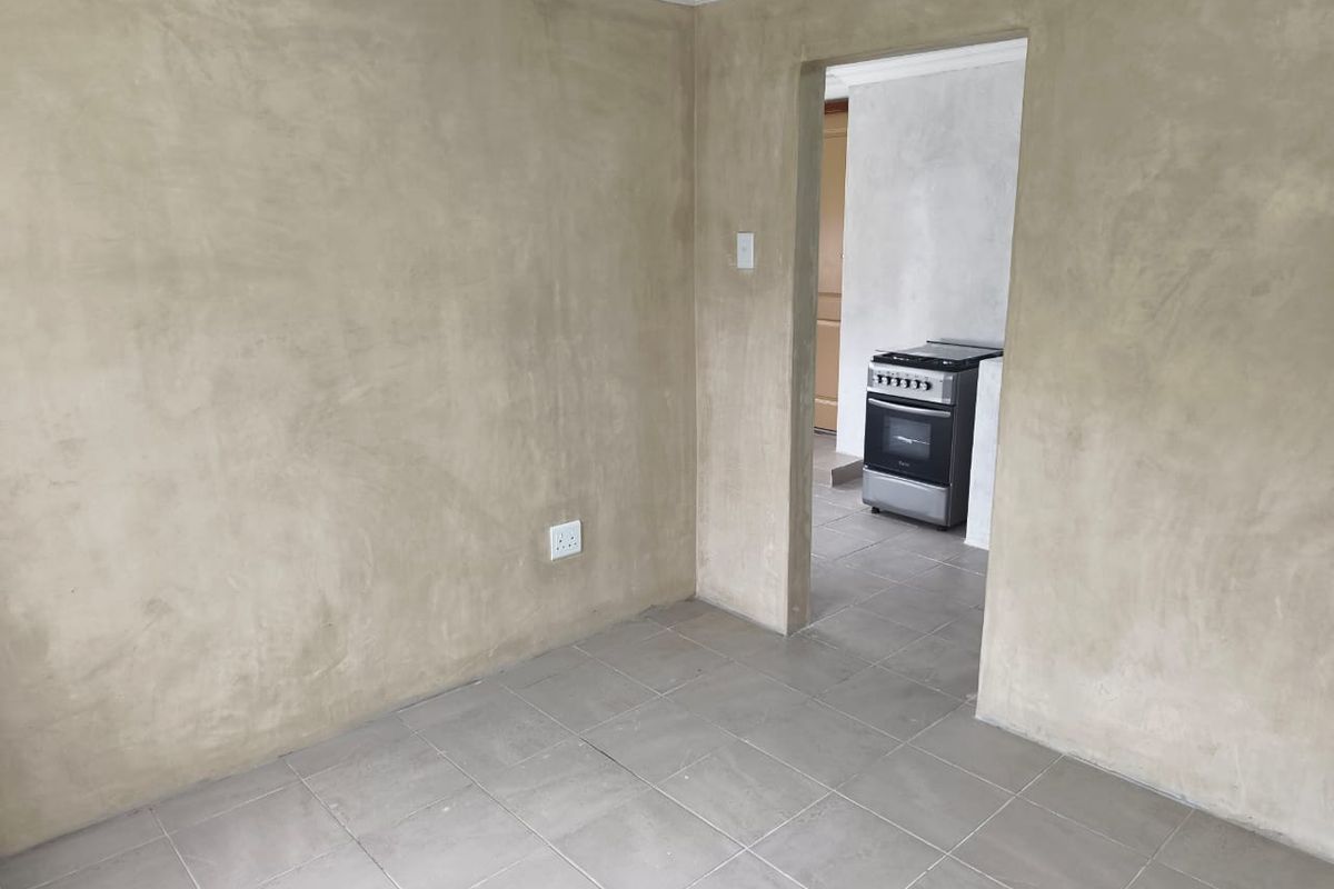 An interior view of a room with unfinished plaster walls and tiled flooring, leading into an adjacent kitchen area featuring a freestanding stove.
