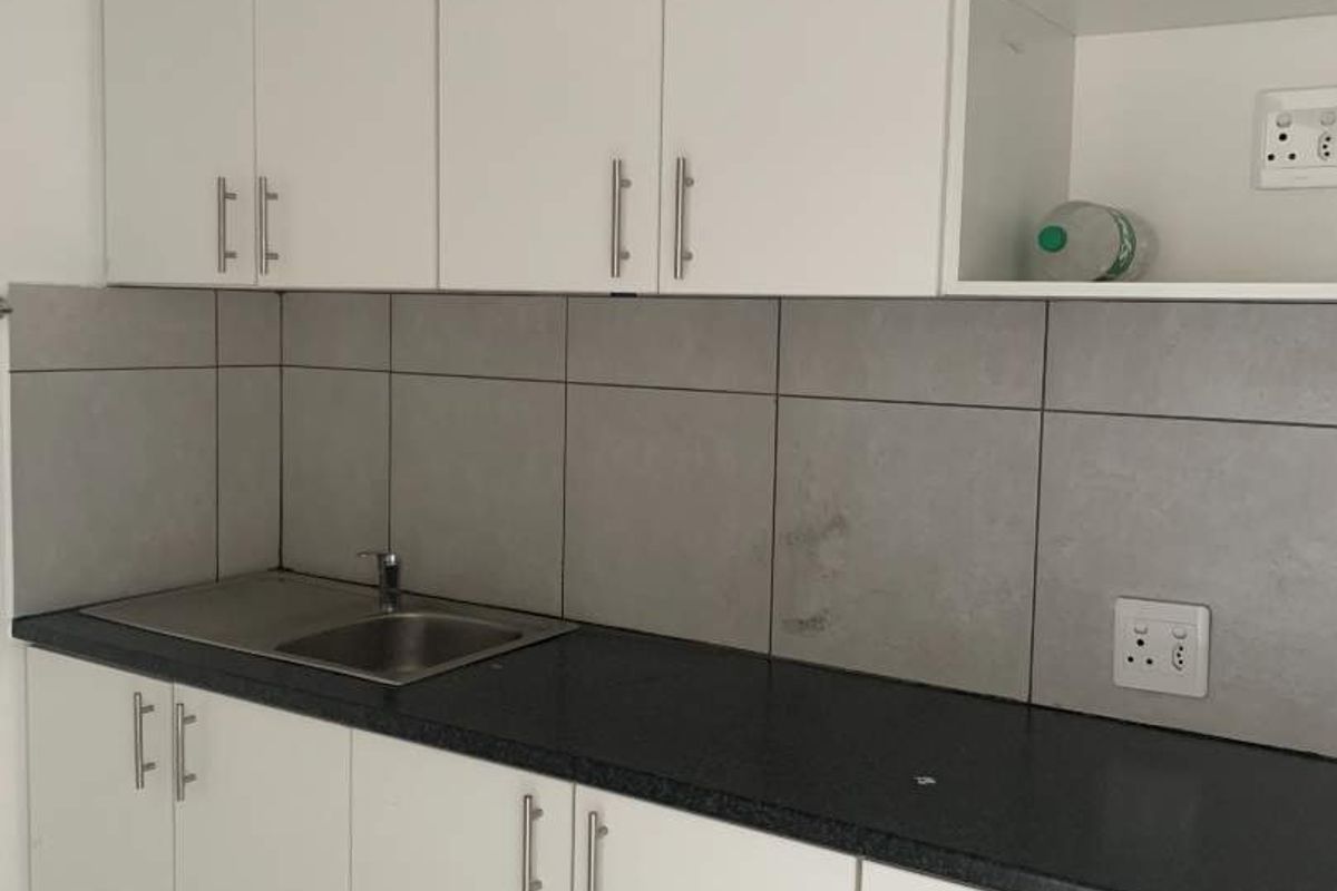 A functional kitchenette featuring white cabinetry, dark laminate countertops, a stainless steel sink, and grey tiled backsplash.