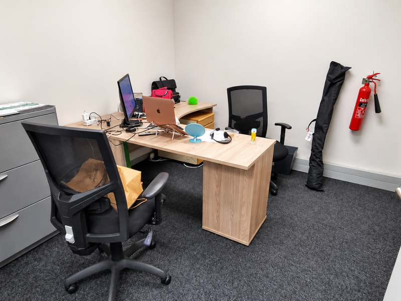 A functional office space featuring carpeted flooring, neutral walls, and a wall-mounted fire extinguisher.