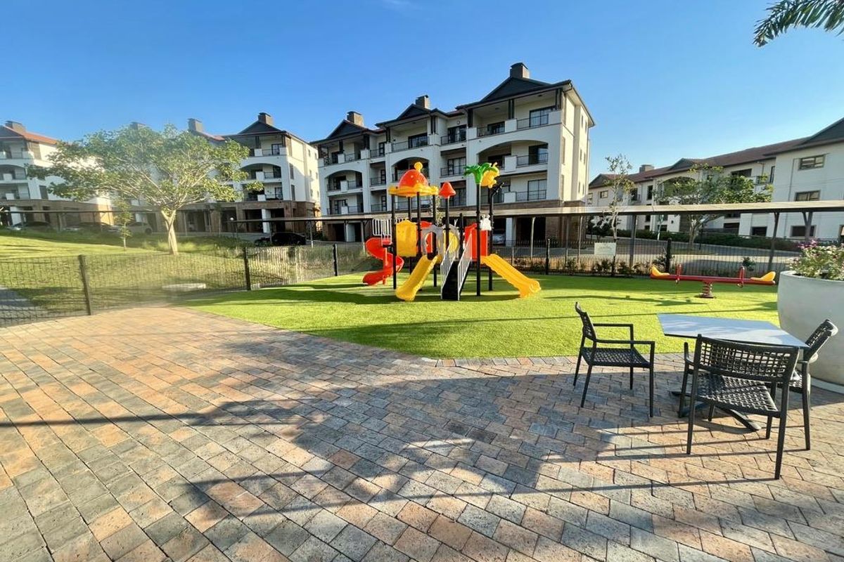 A well-maintained communal outdoor area featuring a paved patio, artificial lawn, and children's playground equipment set against a modern apartment complex.