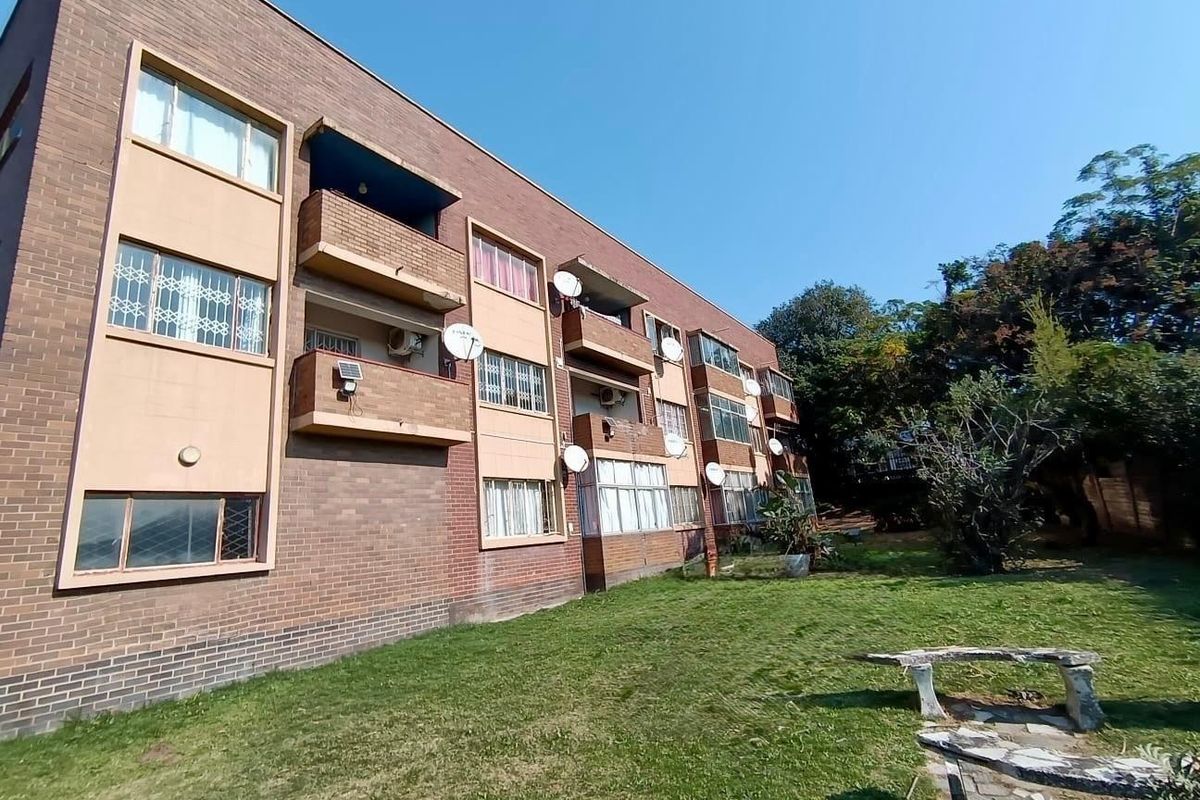 Rear view of a multi-story brick apartment building featuring private balconies, security bars on windows, and established communal garden space.
