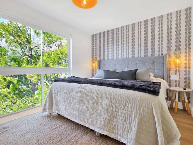 A bright and well-maintained bedroom featuring a large window with leafy views, patterned feature wallpaper, and warm hardwood flooring.