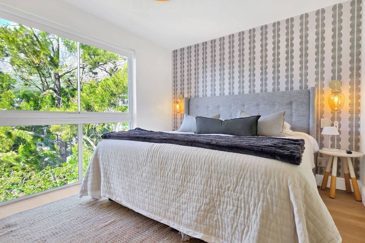 A bright and well-maintained bedroom featuring a large window with leafy views, patterned feature wallpaper, and warm hardwood flooring.