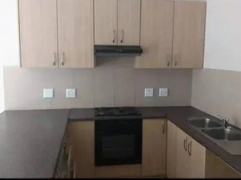 A functional kitchen featuring light wood-finish cabinetry, dark laminate countertops, a built-in oven with hob, and a double stainless steel sink.