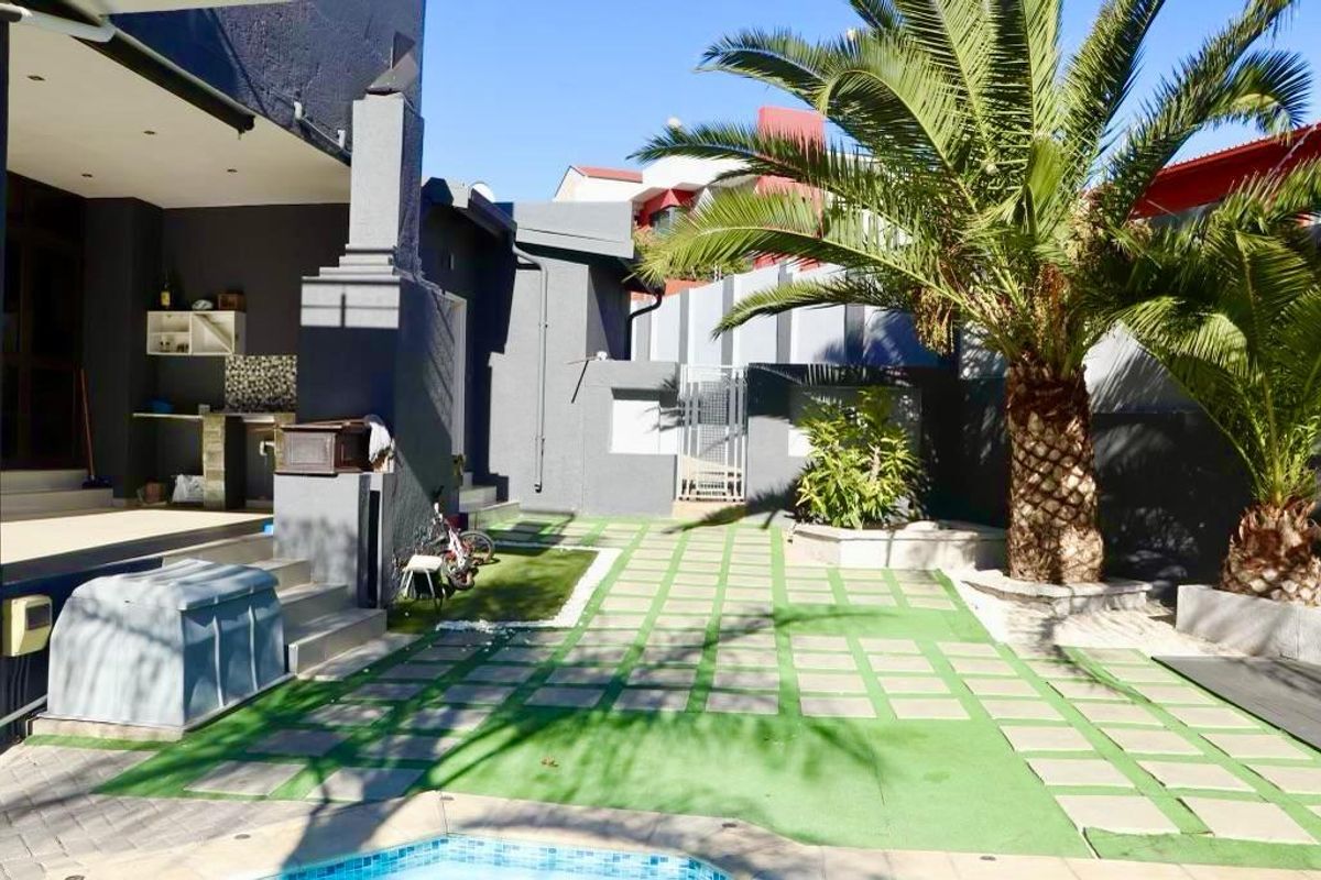 A rear outdoor area featuring a paved patio, built-in braai area, and a swimming pool, surrounded by a landscaped garden with a palm tree.