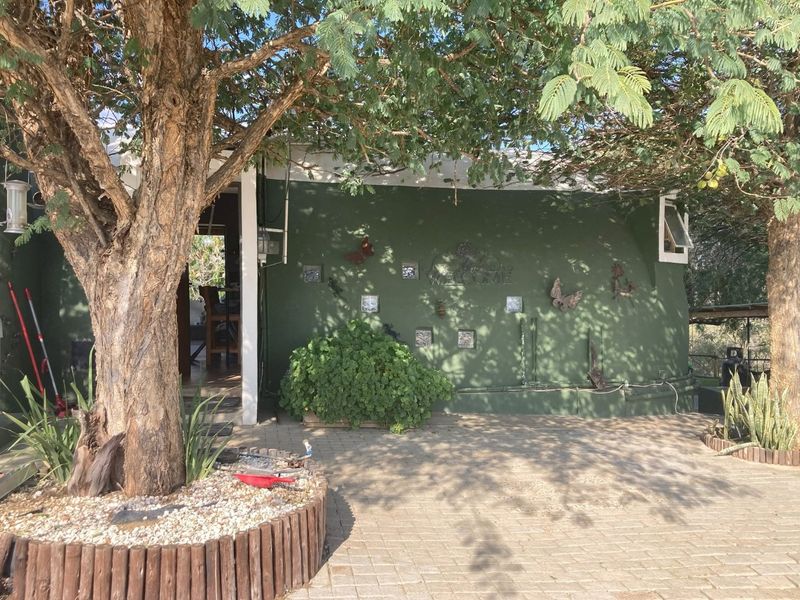 Front exterior view of the house featuring a paved driveway, mature trees, and a green painted facade with decorative wall elements.