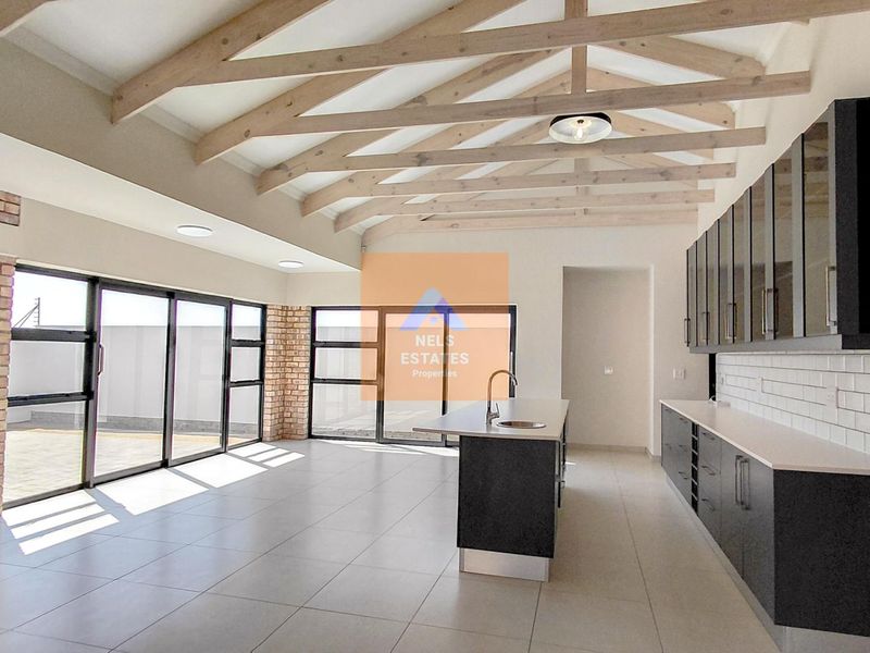 Spacious open-plan living and kitchen area featuring exposed timber beams, a built-in brick fireplace, modern cabinetry, and large sliding doors leading outdoors.