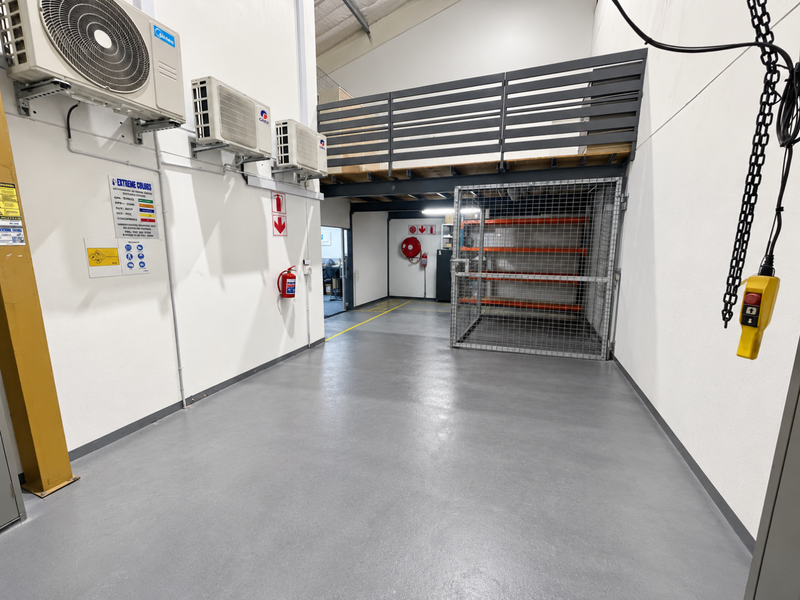 A clean, well-maintained industrial utility space featuring epoxy flooring, a mezzanine level with safety railing, security cage, and wall-mounted air conditioning units.